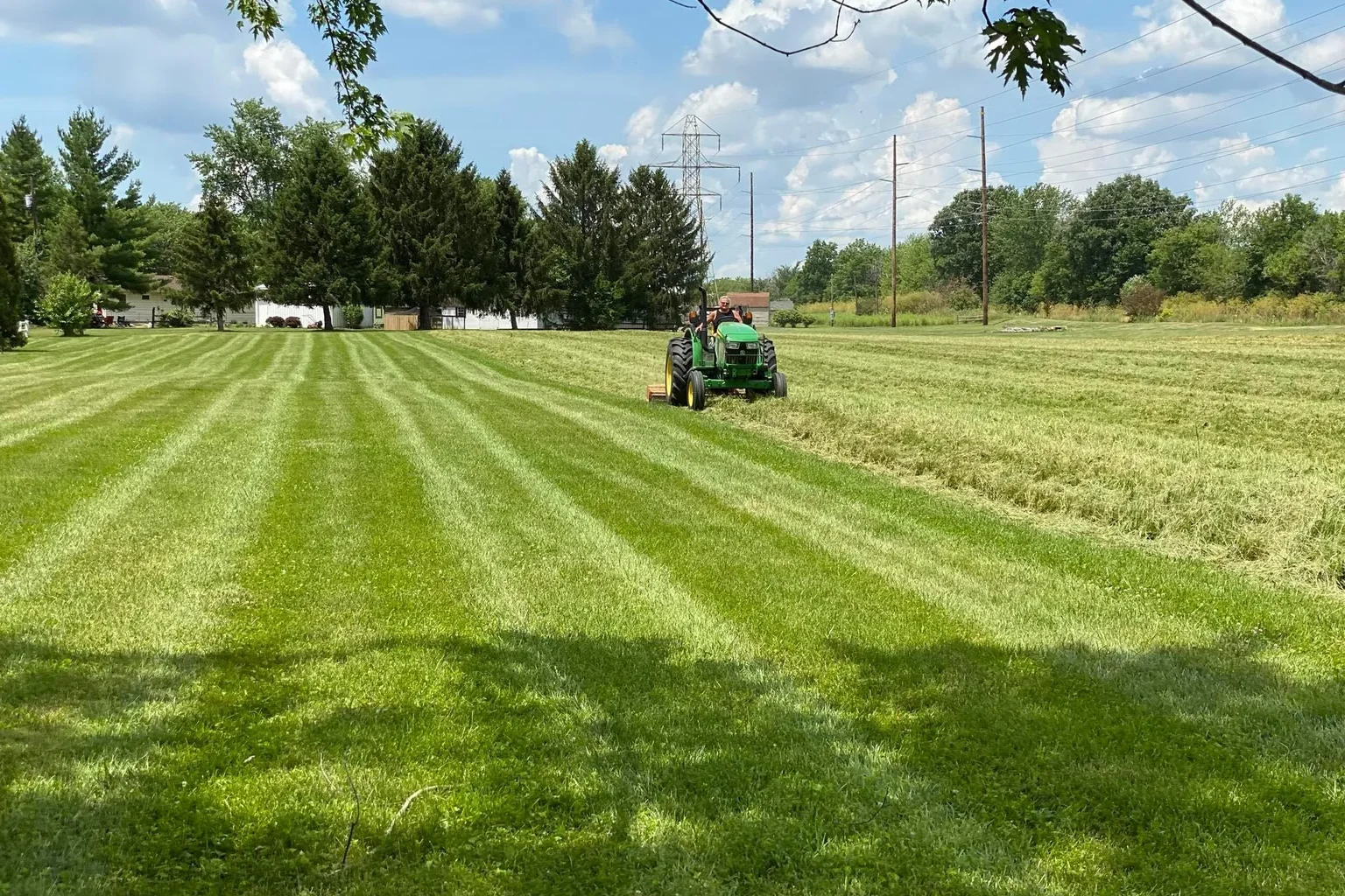 Tractor mowing broad strips of grass beside a tree-lined property